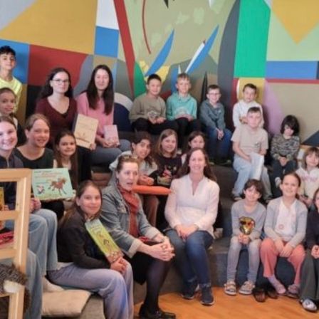 Eine Gruppe von Kindern und Erwachsenen posiert für ein Foto in einem Raum mit bunter Wandkunst. Einige Kinder halten Bücher und Auszeichnungen.