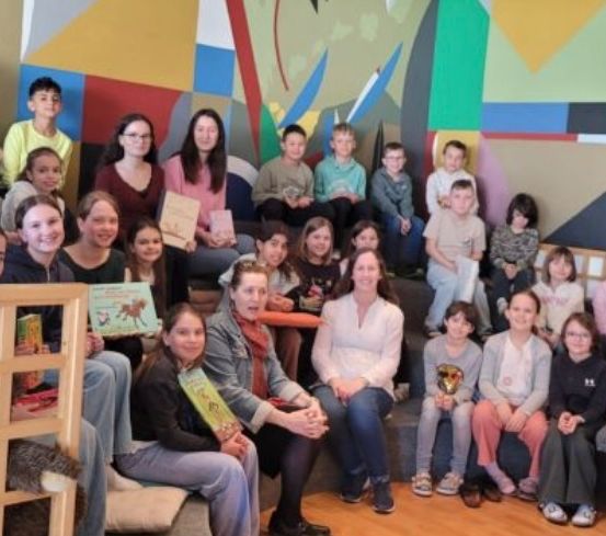 Eine Gruppe von Kindern und Erwachsenen posiert für ein Foto in einem Raum mit bunter Wandkunst. Einige Kinder halten Bücher und Auszeichnungen.