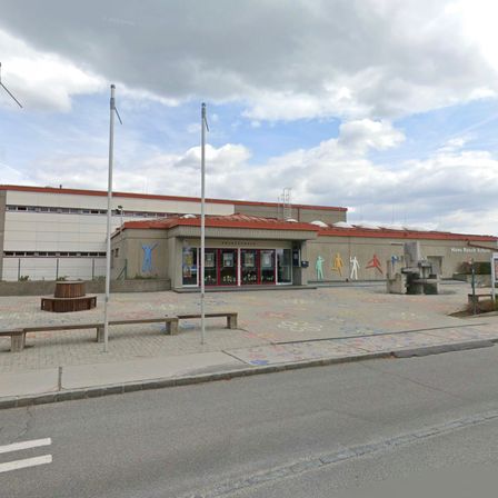 Ein Gebäude mit rotem Dach und einer Menschenmalerei steht unter einem bewölkten Himmel. Davor befindet sich ein Rasenbereich mit Bänken und einer Zeichnung.