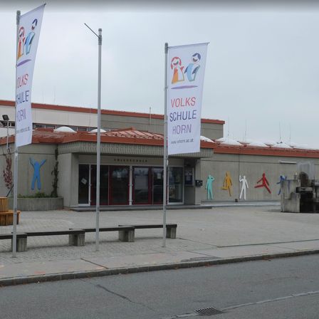 Ein Volksschule Gebäude mit Bannern und bunten Figuren an der Wand. Bänke und eine kleine Holzbank stehen davor.
