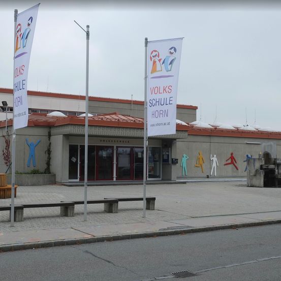 Ein Volksschule Gebäude mit Bannern und bunten Figuren an der Wand. Bänke und eine kleine Holzbank stehen davor.