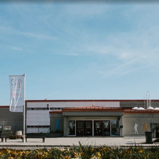 Ein Gebäude mit roten Dächern und weißen Wänden steht vor einem blauen Himmel. Eine Flagge mit blauen und weißen Figuren ist auf einem Pfahl. Es gibt Menschen an der Wand. Ein Mülleimer steht an der Seite.