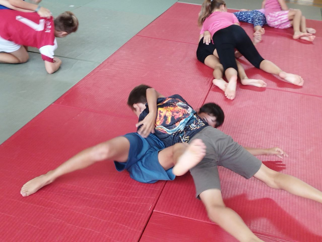 Several children are practicing martial arts on a red mat, with one child lying on the ground and another on top of him.
