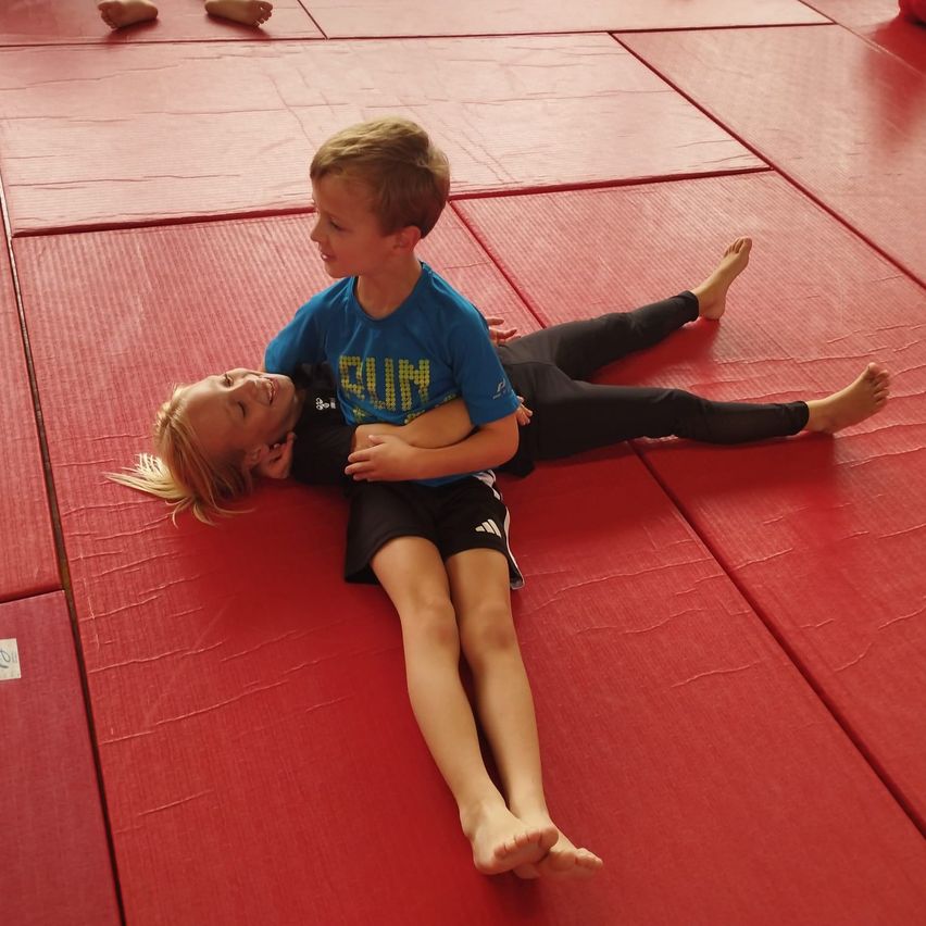 Two children, a boy and a girl, are lying on a red mat, likely practicing martial arts. The boy is wearing a blue shirt with the word 'RUN' on it.