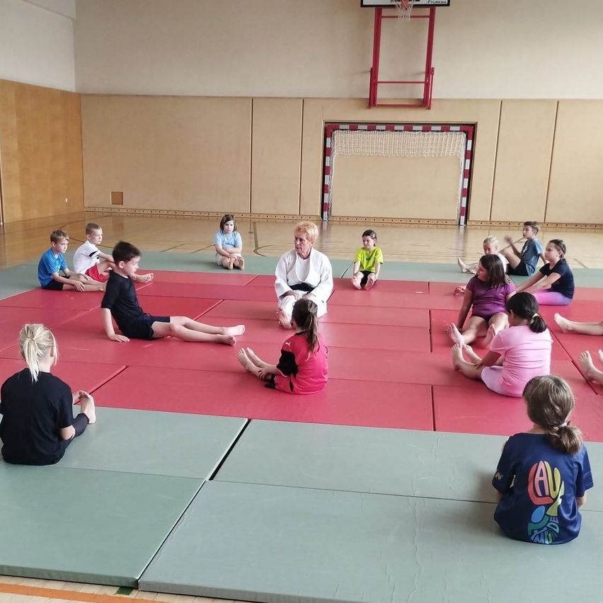 A group of children and an adult are sitting on a red mat in a gym, possibly doing yoga.