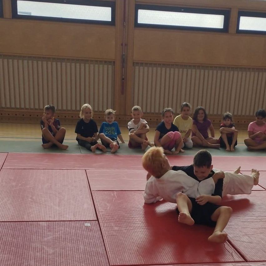 A group of children are sitting in a row watching two boys wrestle on a red mat.