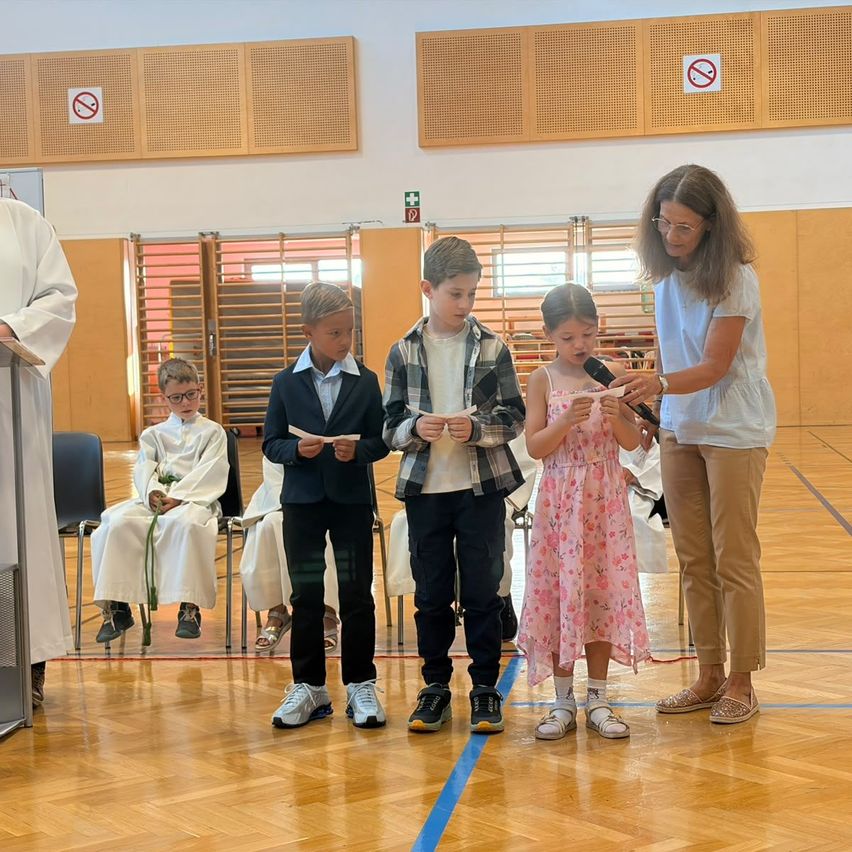 Four children and an adult woman are standing in a room, possibly a church, holding papers and listening to a woman speaking into a microphone.