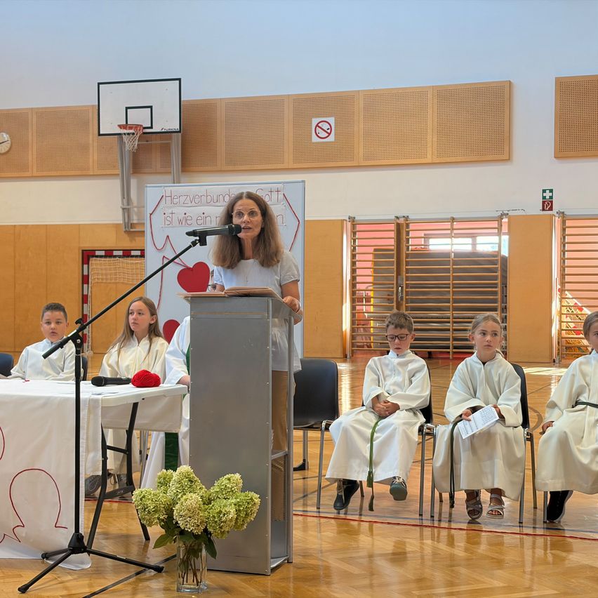 A woman speaks at a podium in a gymnasium, surrounded by children in white robes. A basketball hoop and a sign are visible in the background.