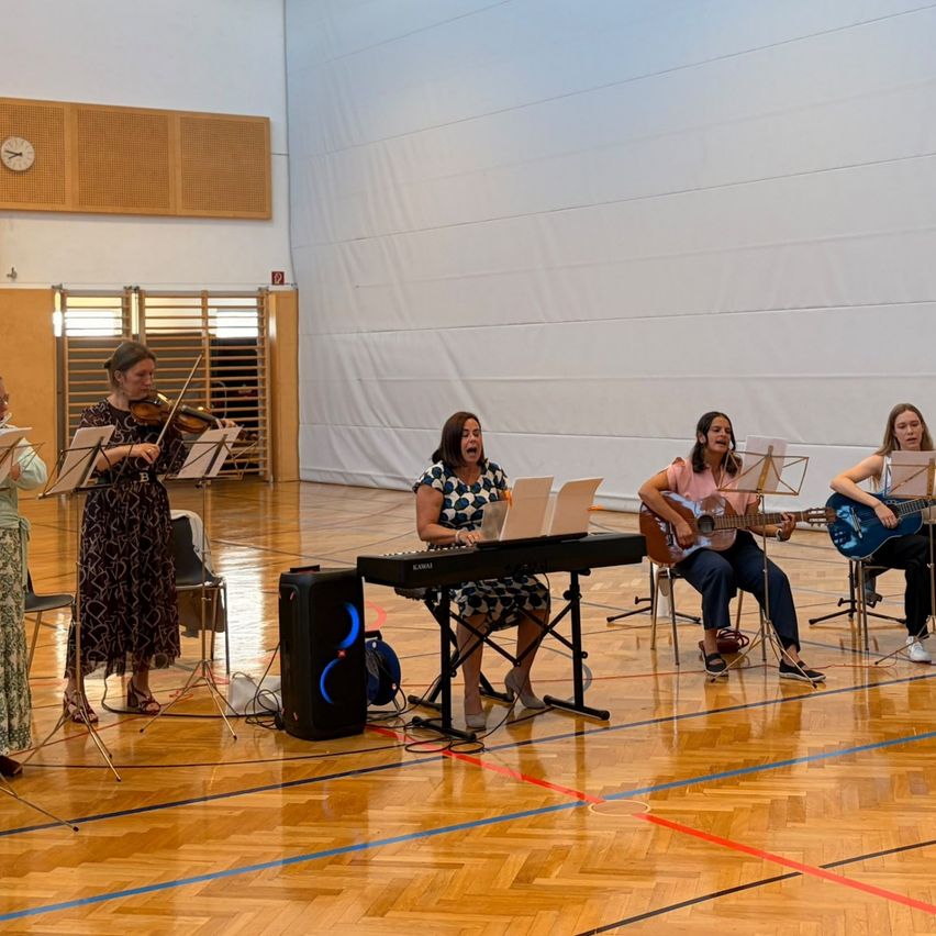 Five women are performing a musical piece in a large room. One plays the violin, another plays the piano, and three others play guitars. They are all singing.