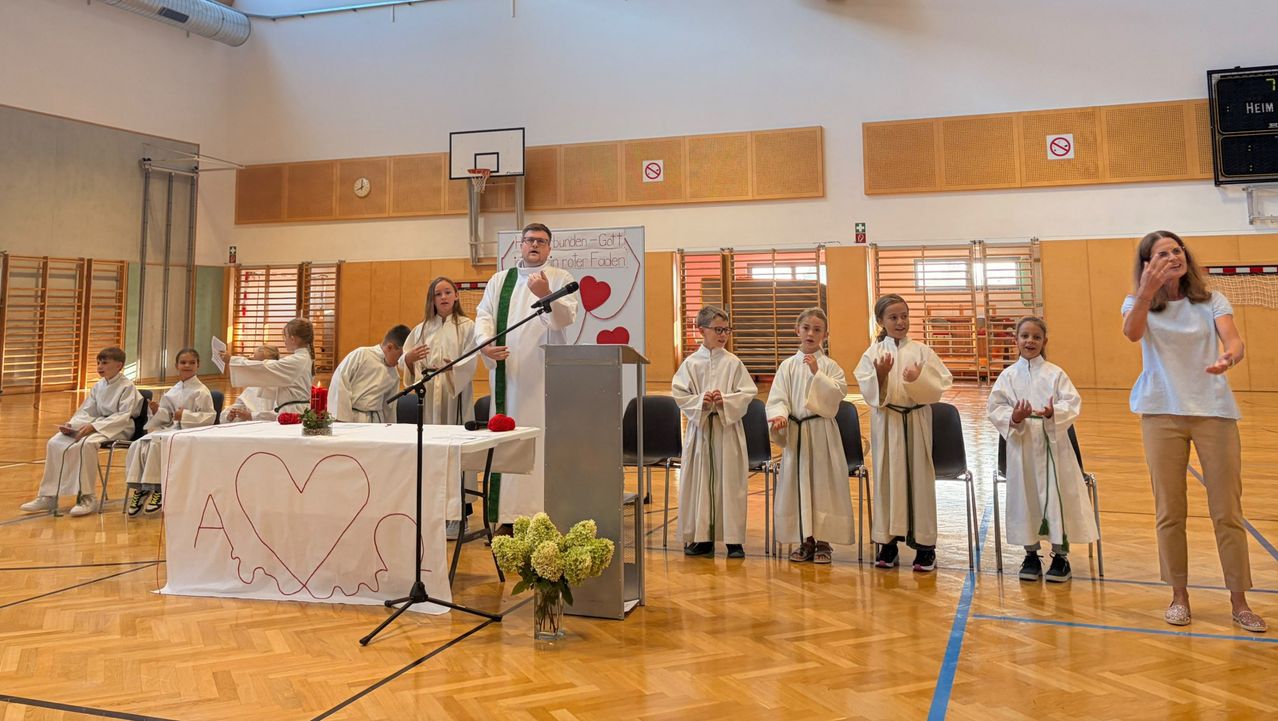 A priest stands at a podium, speaking into a microphone. A group of children in white robes stand nearby, listening attentively. A heart-shaped table and flowers add to the scene.