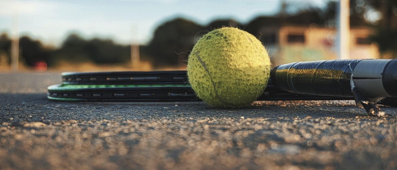 Ein Tennisball liegt neben einem Tennisschläger auf dem Boden. Im Hintergrund ist ein leicht bewölkter Himmel und Bäume zu sehen.
