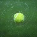 Ein Tennisball in der Luft, aufgenommen mit Wasserspritzern darum herum, vor grünem Hintergrund.