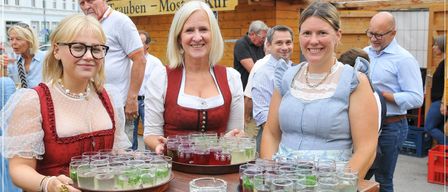Drei Frauen in traditioneller bayerischer Kleidung stehen hinter einem Tisch mit Biergläsern und lächeln in die Kamera. Dahinter ist eine Holzstruktur mit einem Schild Trauben Most Kur zu sehen, mit mehreren Personen im Hintergrund.