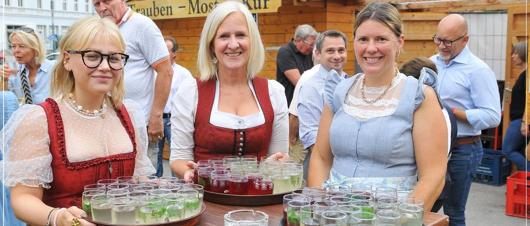 Drei Frauen in traditioneller bayerischer Kleidung stehen hinter einem Tisch mit Biergläsern und lächeln in die Kamera. Dahinter ist eine Holzstruktur mit einem Schild Trauben Most Kur zu sehen, mit mehreren Personen im Hintergrund.