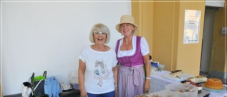 Zwei Frauen stehen hinter einem Tisch mit Tabletts voller Gebäck und Desserts, eine in traditioneller deutscher Kleidung und die andere in lässiger Kleidung.