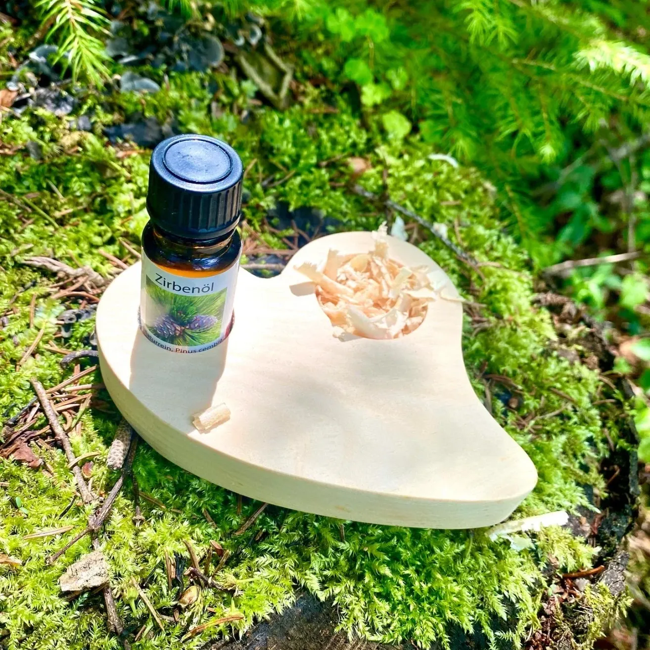 Eine kleine Flasche Zirbenöl steht auf einem holzfarbenen herzförmigen Halter, umgeben von Moos und Holzspänen auf einem Waldboden.