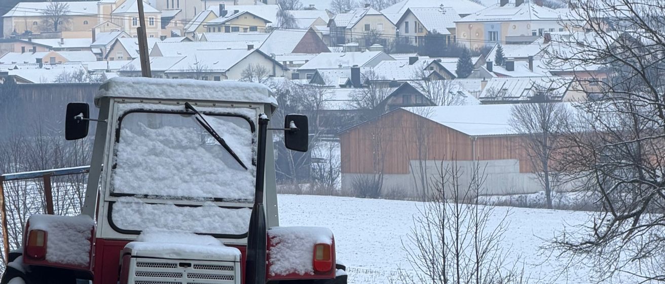 Ein Traktor, bedeckt mit Schnee, steht auf einem verschneiten Feld. Im Hintergrund sind Häuser und eine Kirche von Schnee bedeckt. Der Himmel ist bedeckt.