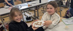 Zwei Mädchen lächeln und halten einen Teller mit Essen. Sie sind in einem Klassenzimmer mit anderen Schülern.