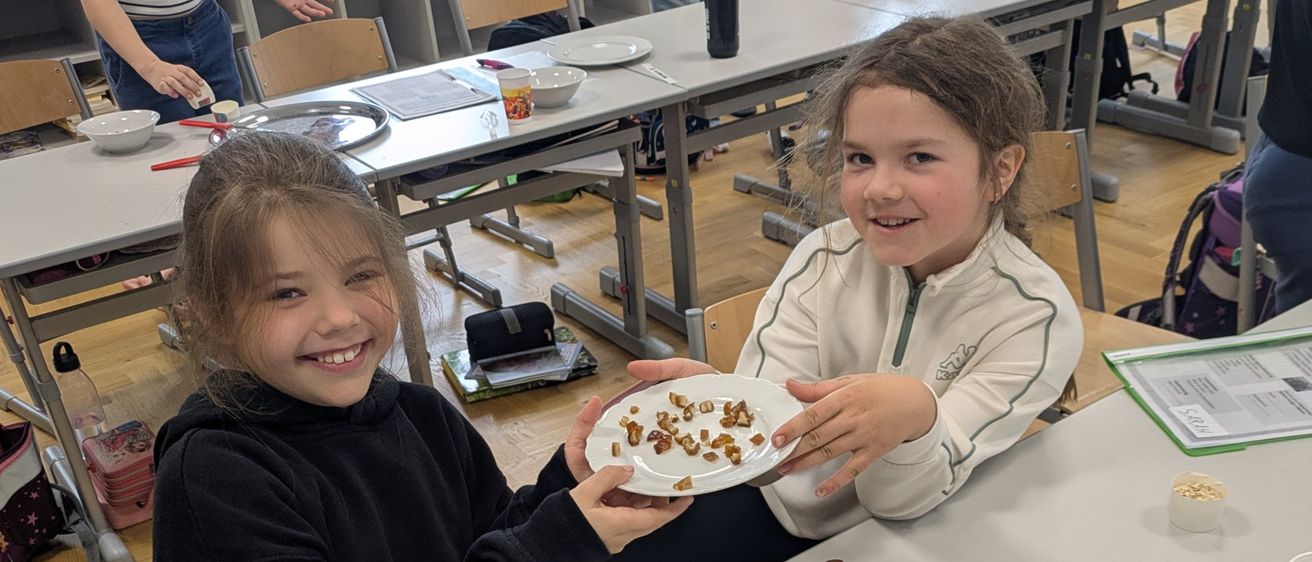 Zwei Mädchen lächeln und halten einen Teller mit Essen. Sie sind in einem Klassenzimmer mit anderen Schülern.