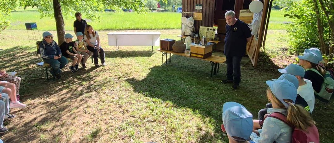 Eine Gruppe von Kindern und Erwachsenen sitzt unter Bäumen und hört einem Mann zu, der in der Nähe einer Holzstruktur mit einem Tisch und Gläsern steht.