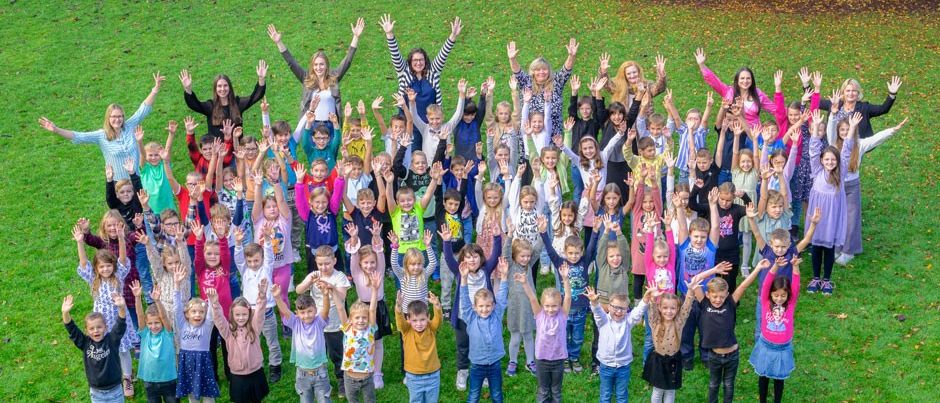 Eine Gruppe von Kindern und ihren Lehrern auf einem Grasfeld, alle mit erhobenen Händen und lächelnd für ein Foto.