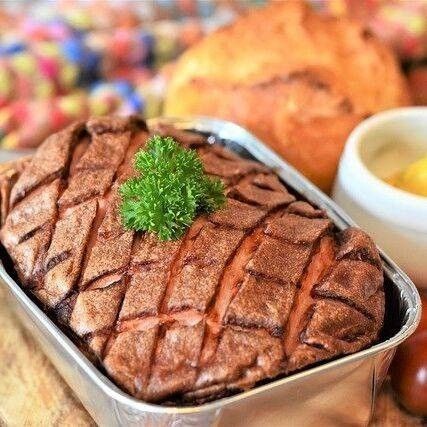 Ein Gericht aus gegrilltem Fleisch mit einem Schnitt Petersilie darauf, serviert in einer silbernen Schale auf einer Holzoberfläche. Dahinter befindet sich eine Schüssel mit Sauce und ein Laib Brot.