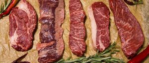 Mehrere Rindfleischstücke sind auf Pergamentpapier angeordnet, mit Rosmarin und grünen Chilis in der Nähe. Das Fleisch variiert in Größe und Form, wobei einige Stücke Marmorierung zeigen.