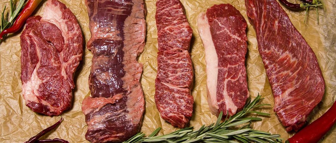 Mehrere Rindfleischstücke sind auf Pergamentpapier angeordnet, mit Rosmarin und grünen Chilis in der Nähe. Das Fleisch variiert in Größe und Form, wobei einige Stücke Marmorierung zeigen.