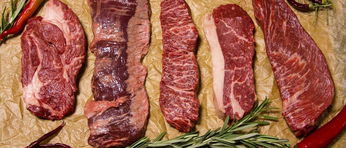 Mehrere Rindfleischstücke sind auf Pergamentpapier angeordnet, mit Rosmarin und grünen Chilis in der Nähe. Das Fleisch variiert in Größe und Form, wobei einige Stücke Marmorierung zeigen.