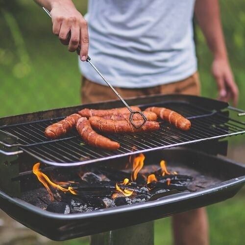 Eine Person steht neben einem Grill, dreht Würste mit einer Zange, über brennende Kohlen und Flammen, in einer Außenumgebung.