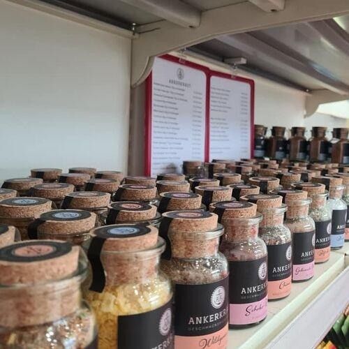 Ein Regal mit verschiedenen kleinen Gläsern mit Korkverschlüssen, beschriftet mit Anker. Die Gläser enthalten verschiedenfarbige Inhalte, möglicherweise Gewürze oder Würzmittel. Über den Gläsern befindet sich ein rotes Schild mit Text.