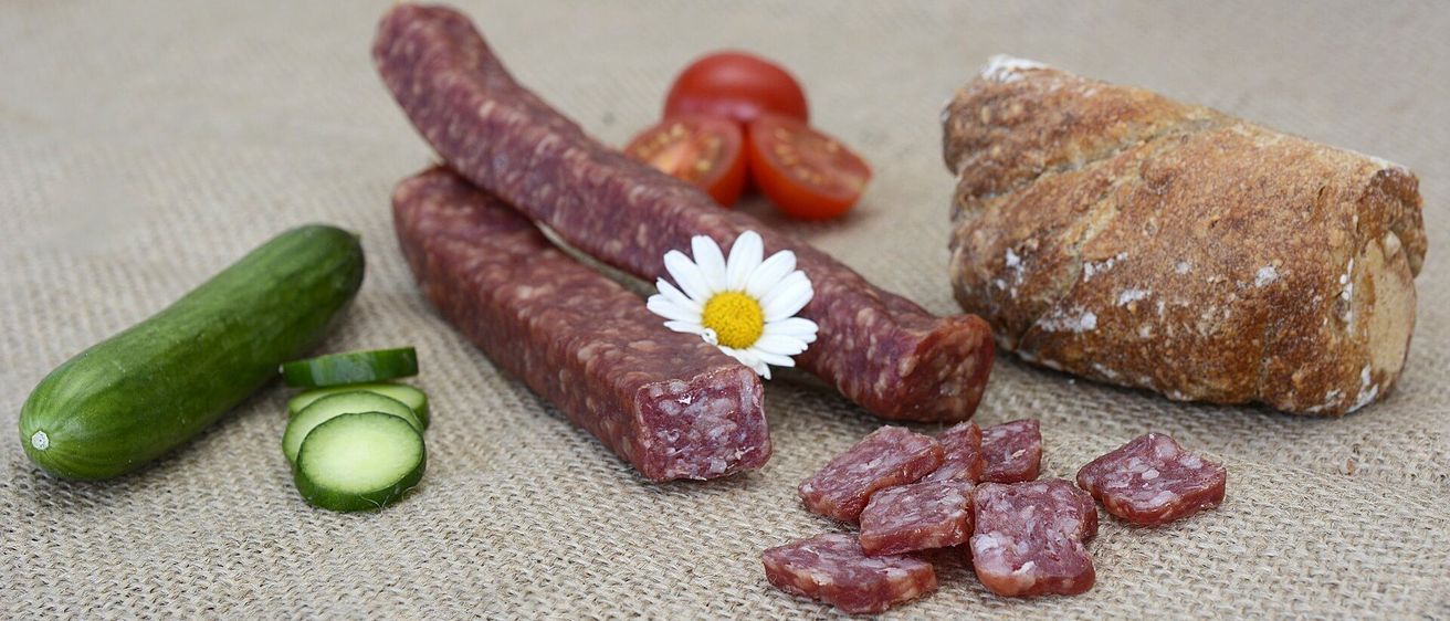 Zwei Würste mit einer Gänseblümchenblüte liegen auf einem Tisch mit Gurken und geschnittenen Tomaten. Im Hintergrund befindet sich ein Stück Brot.