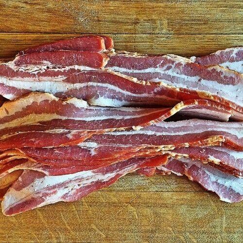 Ein Stapel frisch geschnittener Speck liegt auf einer Holzoberfläche. Der Speck hat eine rosa-rote Farbe mit weißen Fettstreifen.