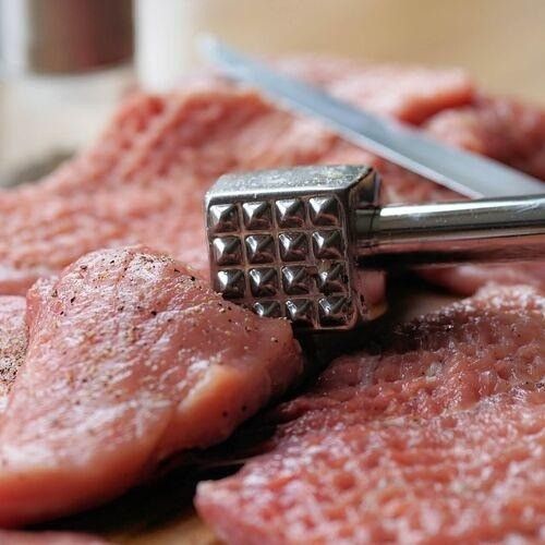 Eine Nahaufnahme von rohem Fleisch auf einem Schneidebrett, mit einem silbernen Fleischklopfer und einem Messer im Vordergrund.