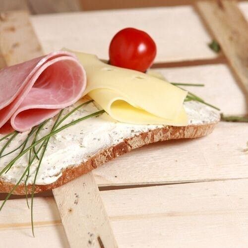 Ein Stück Brot, belegt mit Käse, Schinken und einer Tomate. Es hat ein paar Schnittlauch darüber. Das Brot liegt auf einer Holzoberfläche.