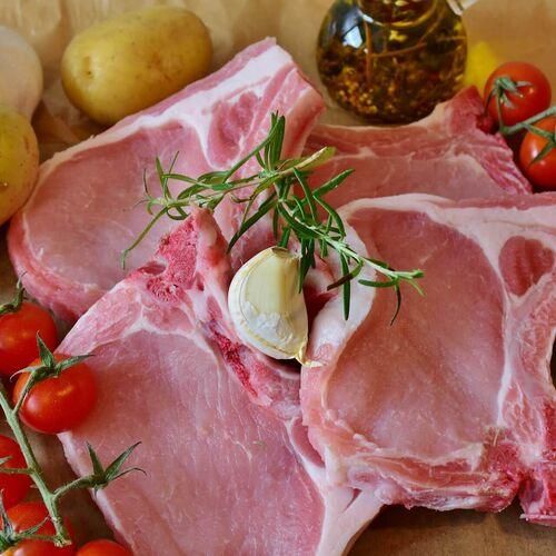 Rohes Schweinefleisch mit Rosmarin und Knoblauch auf einem Holztisch mit Kartoffeln, Tomaten und Öl.