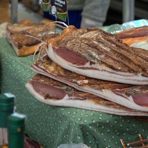Mehrere Scheiben von luftgetrocknetem Fleisch, möglicherweise Speck, sind auf einem mit einem grünen Tuch bedeckten Tisch gestapelt, zusammen mit einer Flasche.