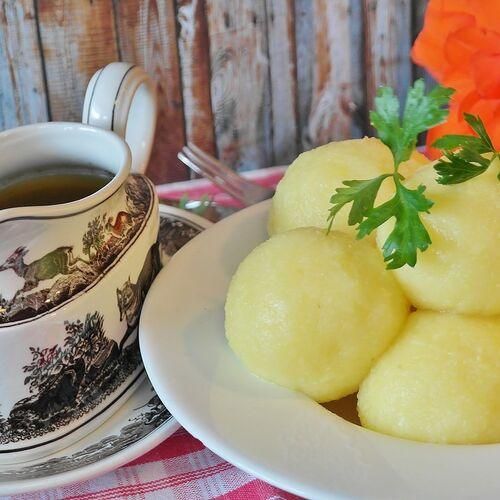 Ein Teller mit gelben Kartoffelklößen, garniert mit Petersilie, steht auf einer karierten Tischdecke. Daneben eine Tasse Tee mit einer blumigen Untertasse. Im Hintergrund ein rustikaler Holzhintergrund.