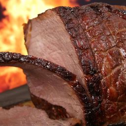 Ein Nahaufnahme eines gebratenen Schinkens mit knuspriger, karamellisiert brauner Kruste, serviert auf einem dunklen Teller vor einem Hintergrund von Flammen.