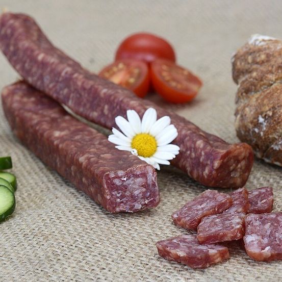 Zwei Stücke Wurst mit einer Gänseblümchenblume auf einem Jutehintergrund, neben Gurken, Tomaten und Brot.