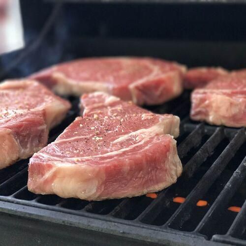 Eine Nahaufnahme mehrerer roher Rindersteaks, die mit Pfeffer und Gewürzen auf einem Grill mit sichtbarer Kohle verfeinert wurden.