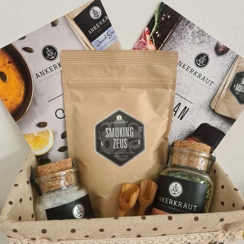 Ein Korb enthält einen Beutel mit Smoking Zeus, ein Glas mit Meersalz, ein Glas mit Kräutern und zwei Holzlöffel. Darüber hinaus befinden sich Broschüren und andere Gegenstände auf dem Korb.
