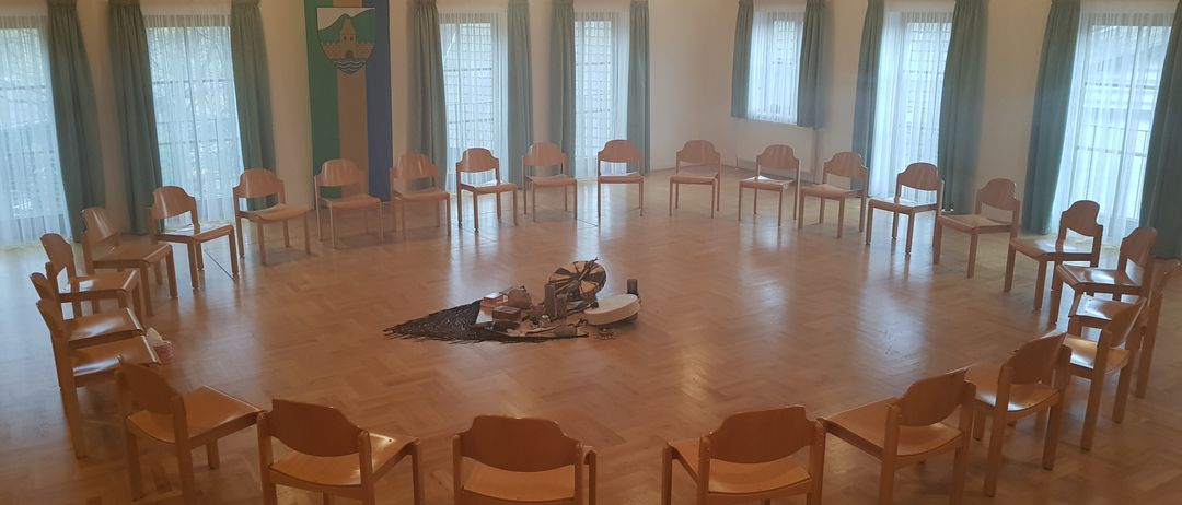 Ein kreisförmiger Raum mit Holzböden und Stühlen, die im Kreis angeordnet sind. In der Mitte befindet sich ein Tisch mit verschiedenen Gegenständen darauf. Der Raum hat große Fenster mit Vorhängen.