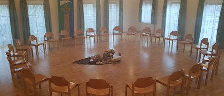 Ein leerer Raum mit Holzboden, mehreren hölzernen Stühlen, die in einem Kreis angeordnet sind, und einigen Gegenständen auf dem Boden. Es gibt Lampen, die von der Decke hängen, Fenster mit Vorhängen und einen Projektor in der Ecke.