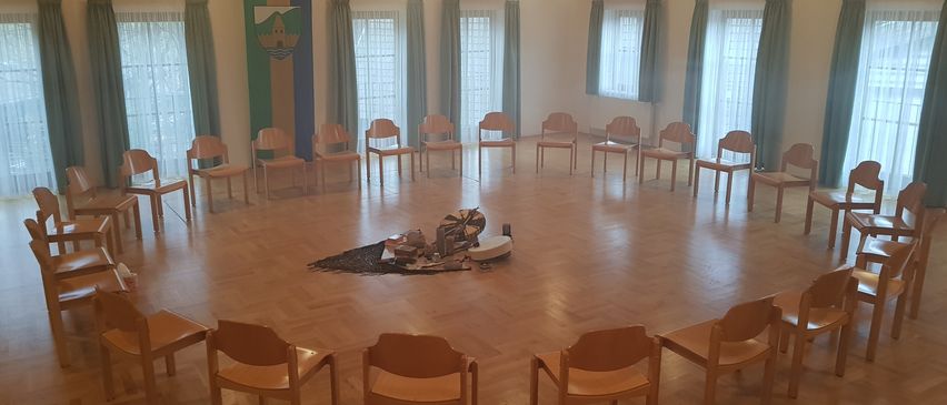 Ein kreisförmiger Raum mit Holzböden und Stühlen, die in einem Kreis angeordnet sind. Ein kleiner Teppich und Gegenstände liegen auf dem Boden. Der Raum hat Fenster mit Vorhängen, einen Projektor und Lampen.