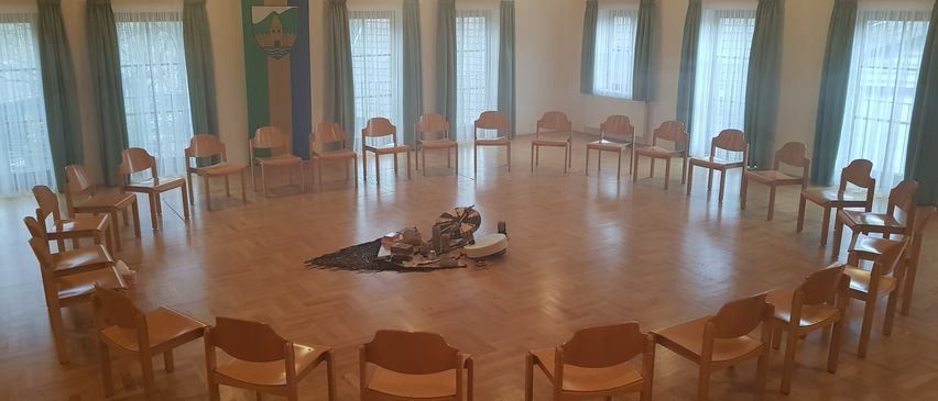 An empty room with wooden floors and several chairs arranged in a circle. A table is in the center, surrounded by various objects. Large windows with curtains are on the walls.