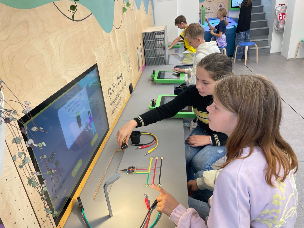 Mehrere Kinder sitzen um einen Tisch herum und beschäftigen sich mit einer praktischen Aktivität mit Kabeln und einem Bildschirm, der eine Comicfigur zeigt.