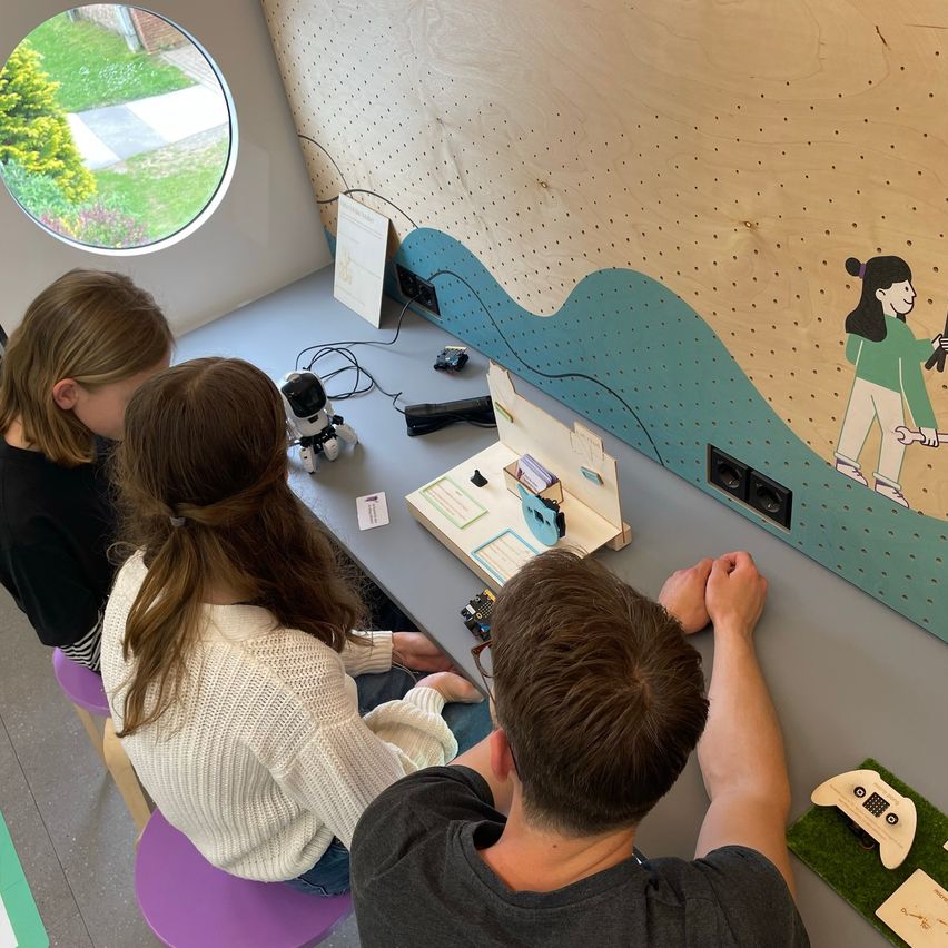 Drei Personen, zwei Frauen und ein Mann, sitzen in einem Raum mit einem runden Fenster. Sie beschäftigen sich mit einer Aktivität, die einen Roboter, ein Buch und andere Gegenstände auf einem Schreibtisch beinhaltet.