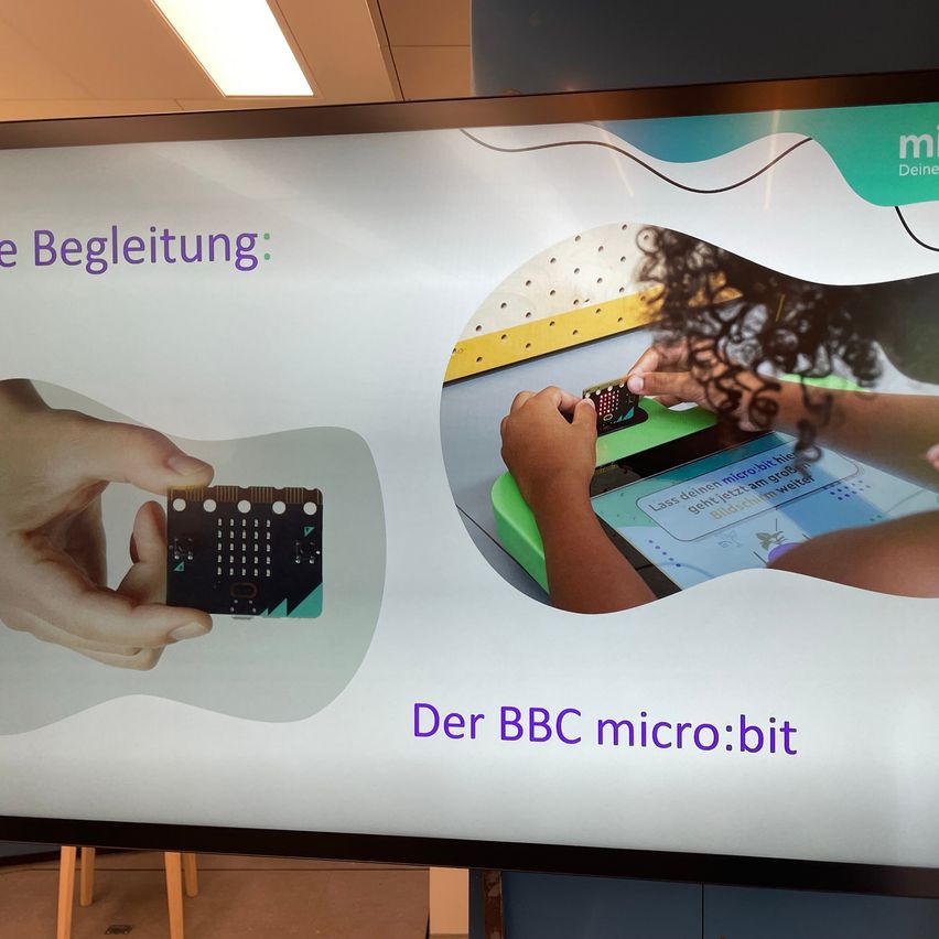 Ein Poster zeigt eine Hand, die ein BBC micro:bit Mikrocontroller hält, eine Person benutzt es. Es steht 'Deine Begleitung: Der BBC micro:bit'.