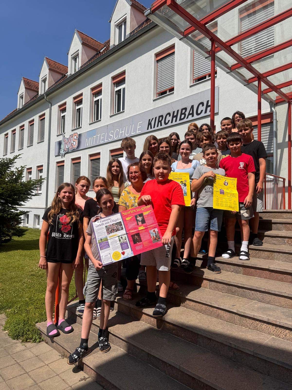 Eine Gruppe junger Menschen steht auf den Stufen vor einem Schulgebäude. Sie halten gelbe Papiere und ein Junge hält ein rotes Papier. Der Name der Schule lautet 'Mittelschule Kirchbach'.